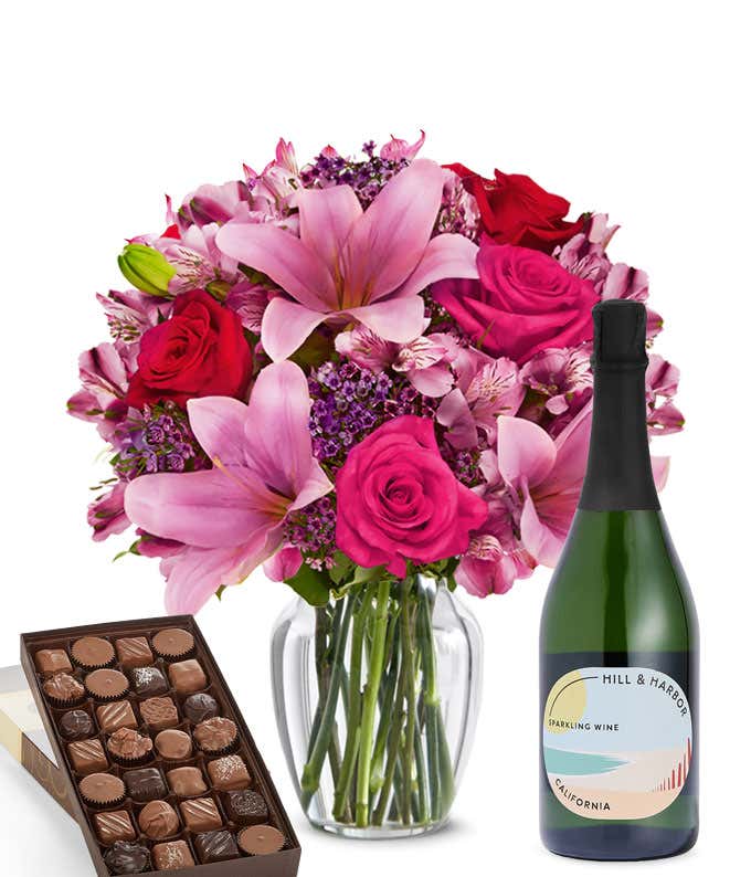 A mixed bouquet of pink and purple flowers in a clear glass vase is paired with a bottle of Hill & Harbor Sparkling Wine and a box of assorted chocolates.