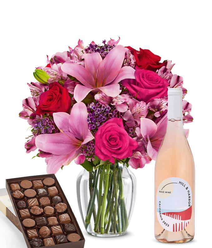 A mixed bouquet of pink and purple flowers in a clear glass vase is paired with a bottle of Hill & Harbor Rosé and a box of assorted chocolates.