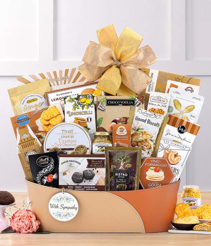 Tan and brown gift basket with a gold bow, filled with assorted snacks including cookies, chocolates, tea, hummus, olives, naan crisps, and cheese spread, displayed on a marble surface with a small flower vase.