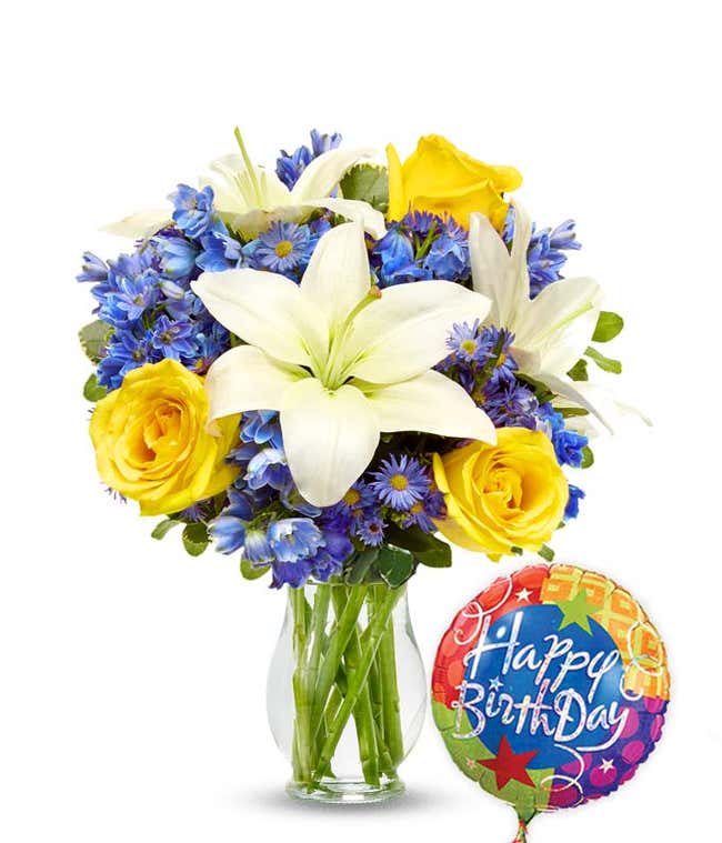 Birthday flower bouquet with white lilies, yellow roses, blue delphinium, and purple asters in a clear glass vase, paired with a colorful Happy Birthday balloon.