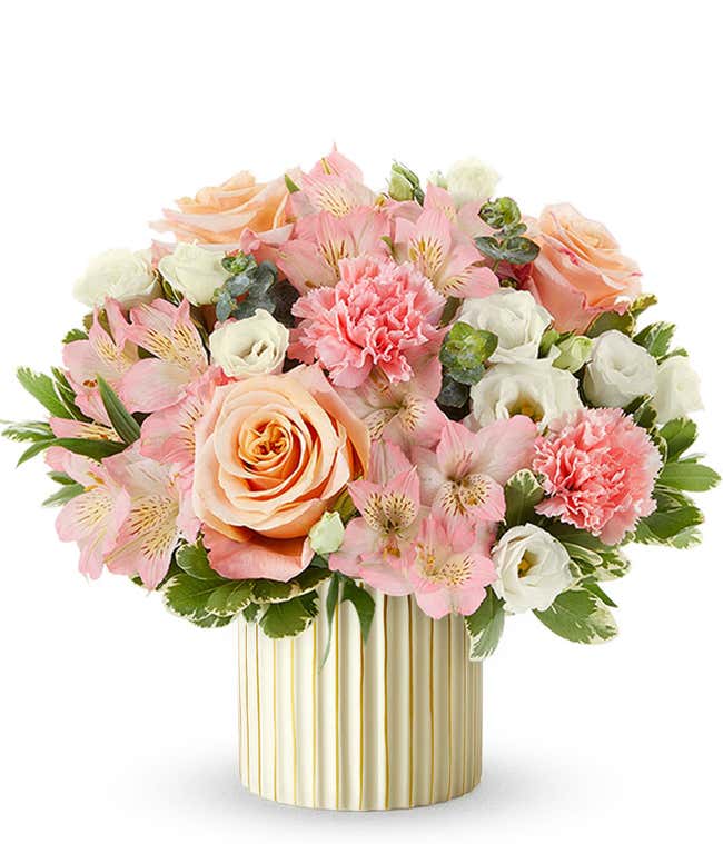 Arrangement of peach roses, pink alstroemeria, pink carnations, white blooms, and variegated greenery in a ribbed ivory planter with gold accents.