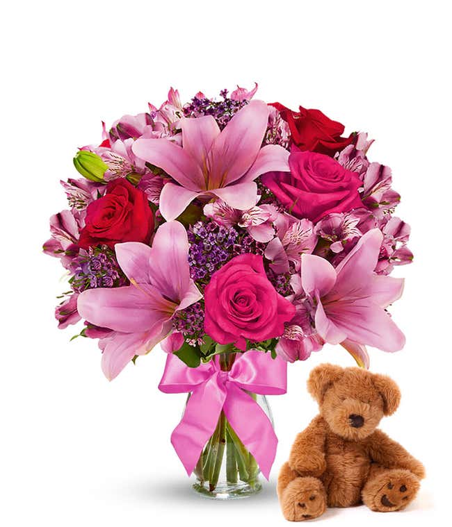 Red and pink roses, pink alstroemeria in a clear vase with bow, accompanied by a keepsake teddy bear.