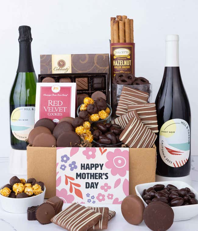 Two bottles of wine - red &amp; sparkling, sit beside chocolate-covered pretzels, mini chocolate chip cookies, caramel popcorn, assorted chocolates, mini buckeyes, chocolate covered Oreos, and a Happy Mother's Day card.
